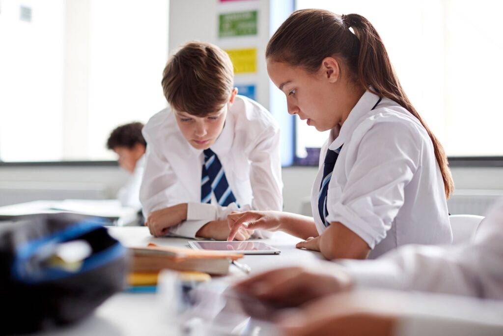 Zwei Schüler in Schuluniform arbeiten konzentriert an einem Tablet in einem Klassenzimmer einer internationalen Privatschule in Berlin, während andere Schüler im Hintergrund lernen.