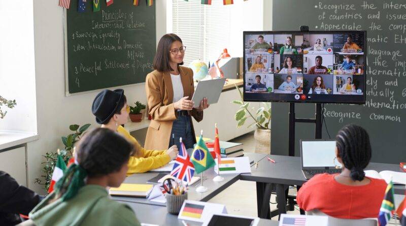 Eine Lehrerin hält in einer internationalen Privatschule in Berlin eine Vorlesung über die Prinzipien internationaler Beziehungen, während Schülerinnen und Schüler über eine Videokonferenz teilnehmen und Flaggen verschiedener Länder auf ihren Tischen stehen.