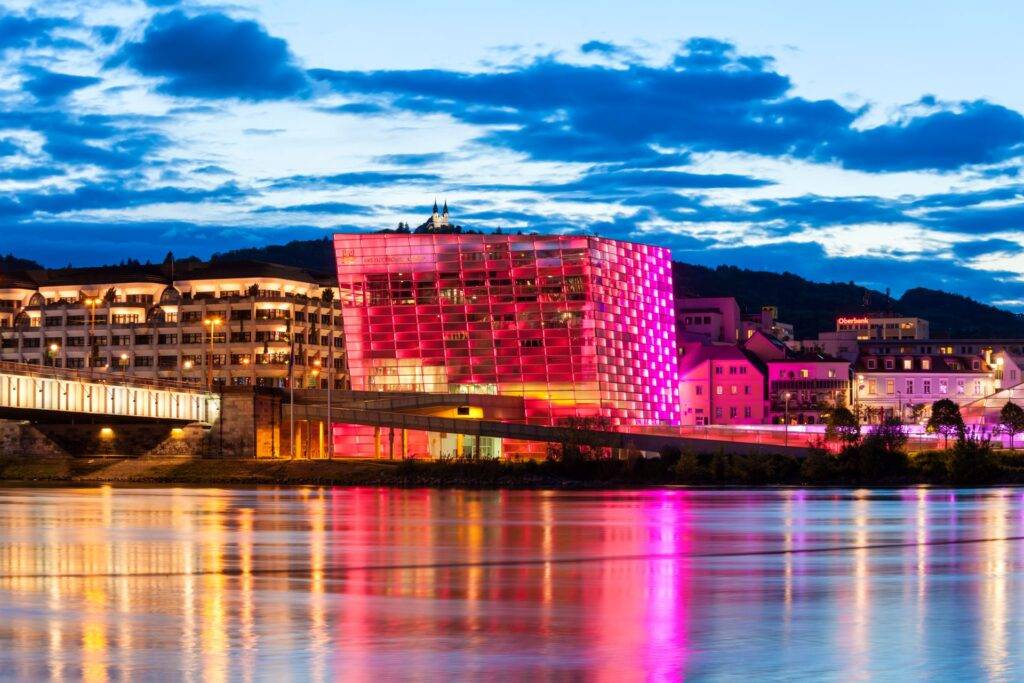 Beleuchtetes Ars Electronica Center in Linz mit pinker Fassade und Spiegelung in der Donau bei Nacht