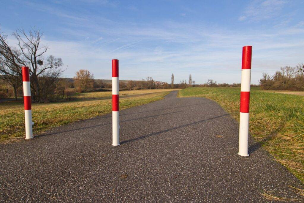 Drei rot-weisse Absperrpfosten sichern einen asphaltierten Weg in freier Landschaft – einfache und effektive Form der Wegbegrenzung