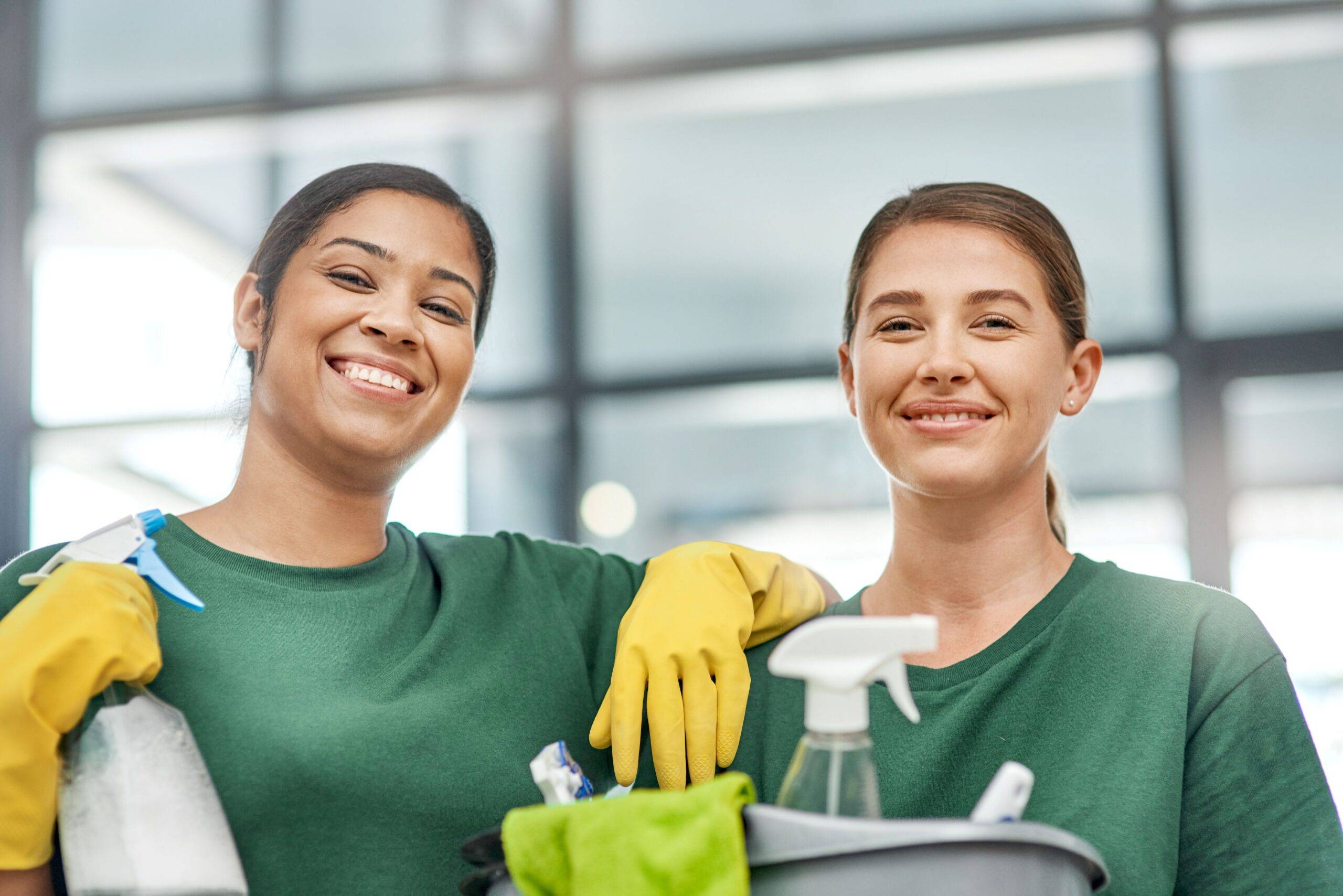 Zwei Reinigungskräfte mit Arbeitskleidung, Handschuhen und Reinigungszubehör. Beide lächeln und wirken professionell sowie freundlich